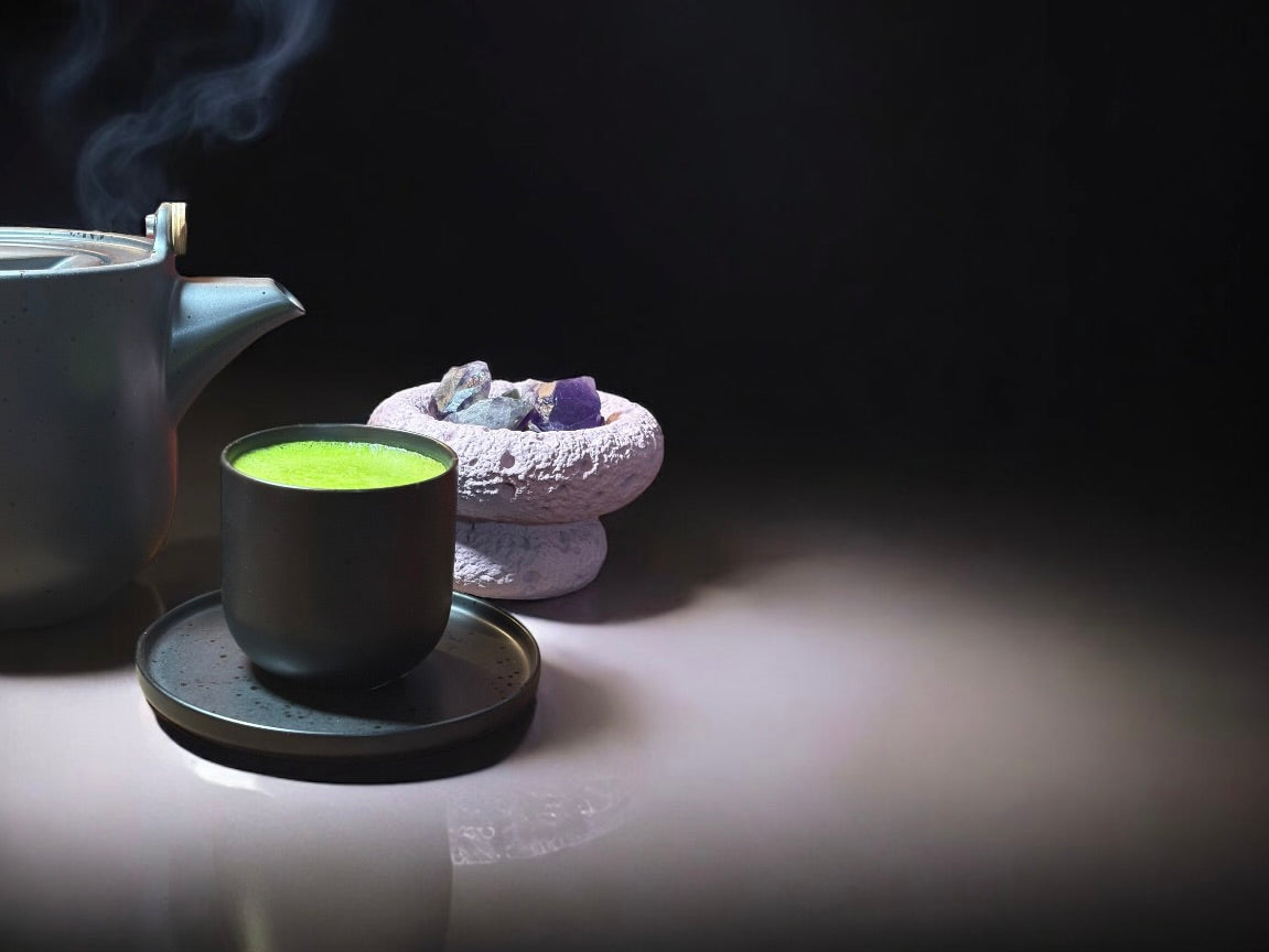 Eine dampfende Teekanne und eine Tasse mit leuchtend grünem Matcha Tee stehen auf einer Untertasse. In der Nähe befindet sich eine runde Steinschale mit mehreren bunten Kristallen, die alle auf einer glatten, spiegelnden Oberfläche vor einem dunklen Hintergrund angeordnet sind.