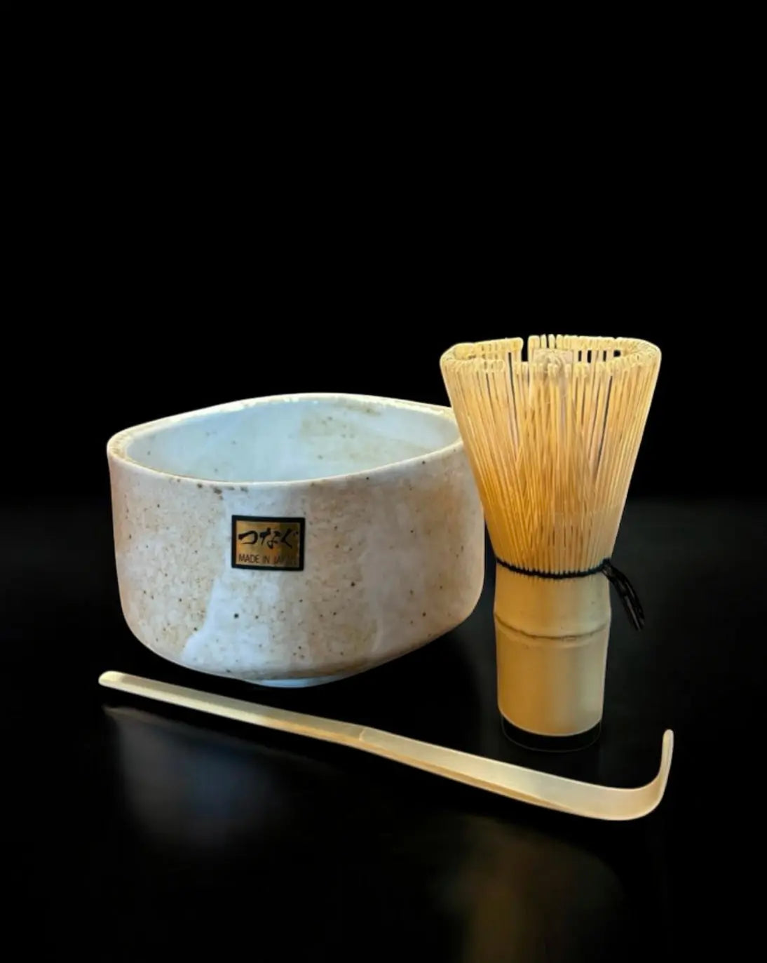 Eine Matcha-Schale aus Keramik, ein Bambusbesen und ein Bambuslöffel sind auf einer spiegelnden Oberfläche mit einem weichen Farbverlauf angeordnet.