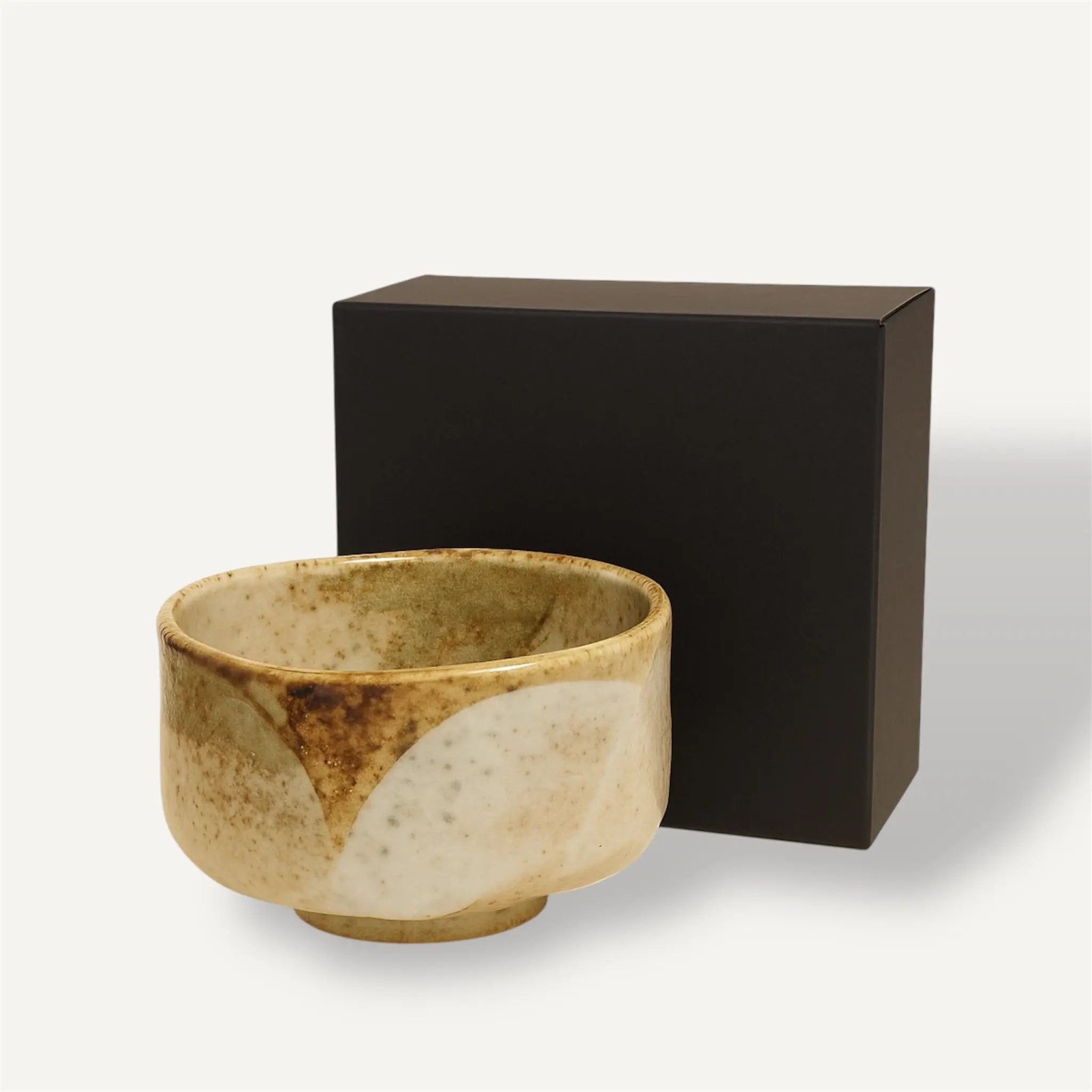 Die Fitaura Matcha Schale Chawan Yuki, handgemacht aus Raku Keramik in Beige und Braun, steht vor einer schlichten schwarzen Box auf weißem Hintergrund.