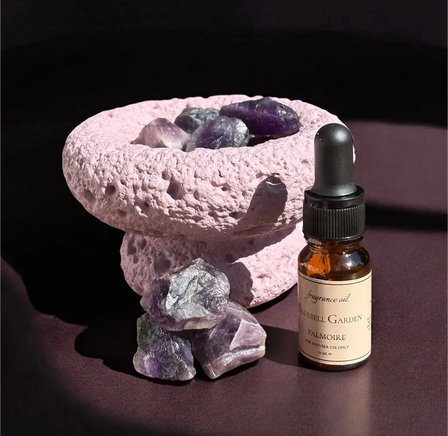 Eine rosafarbene, strukturierte Schale enthält Amethyst-Kristalle, daneben weitere. Daneben steht der Fitaura Lila Mond Aromatherapie-Diffusor mit Amethyst - Zeder & Adlerholz, alles auf einer dunklen Fläche.