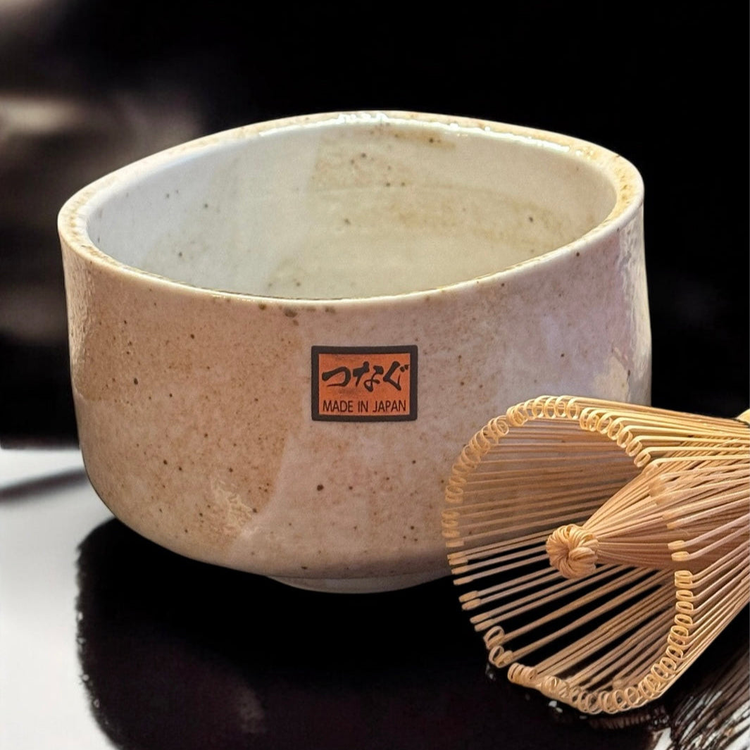 Eine strukturierte beigefarbene japanische Teeschale aus Keramik steht neben dem Fitaura Matchabesen Chasen aus weißem Bambus mit 100 Borsten auf einer glänzenden schwarzen Oberfläche. Die Schale trägt ein Etikett "Made in Japan" mit japanischen Schriftzeichen darüber.