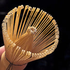 Nahaufnahme des Fitaura Matchabesen Chasen aus weißem Bambus mit 100 Borsten, der seine dünnen, gebogenen Zinken für die Matcha-Zubereitung in der japanischen Teezeremonie auffächert, vor einem dunklen Hintergrund.
