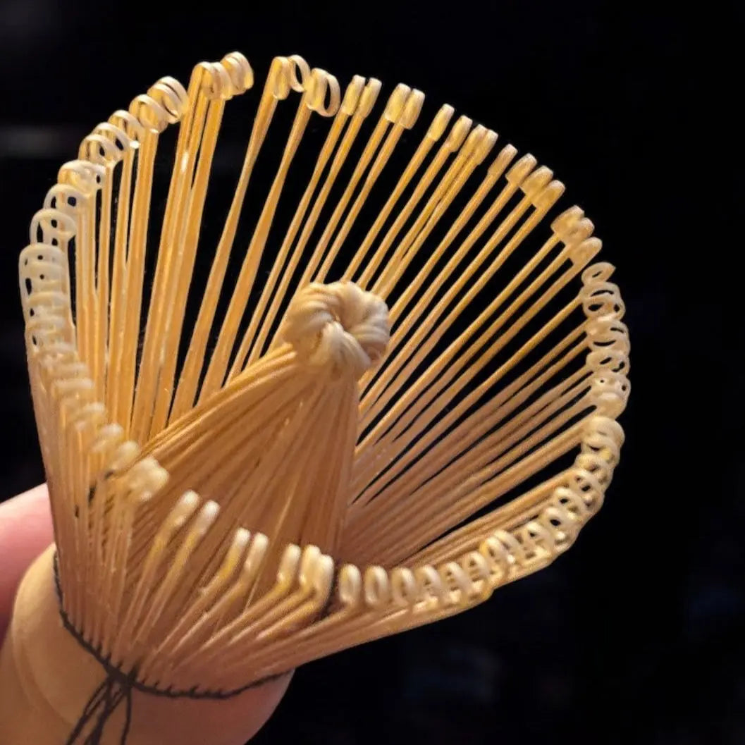 Nahaufnahme des Fitaura Matchabesen Chasen aus weißem Bambus mit 100 Borsten, der seine dünnen, gebogenen Zinken für die Matcha-Zubereitung in der japanischen Teezeremonie auffächert, vor einem dunklen Hintergrund.