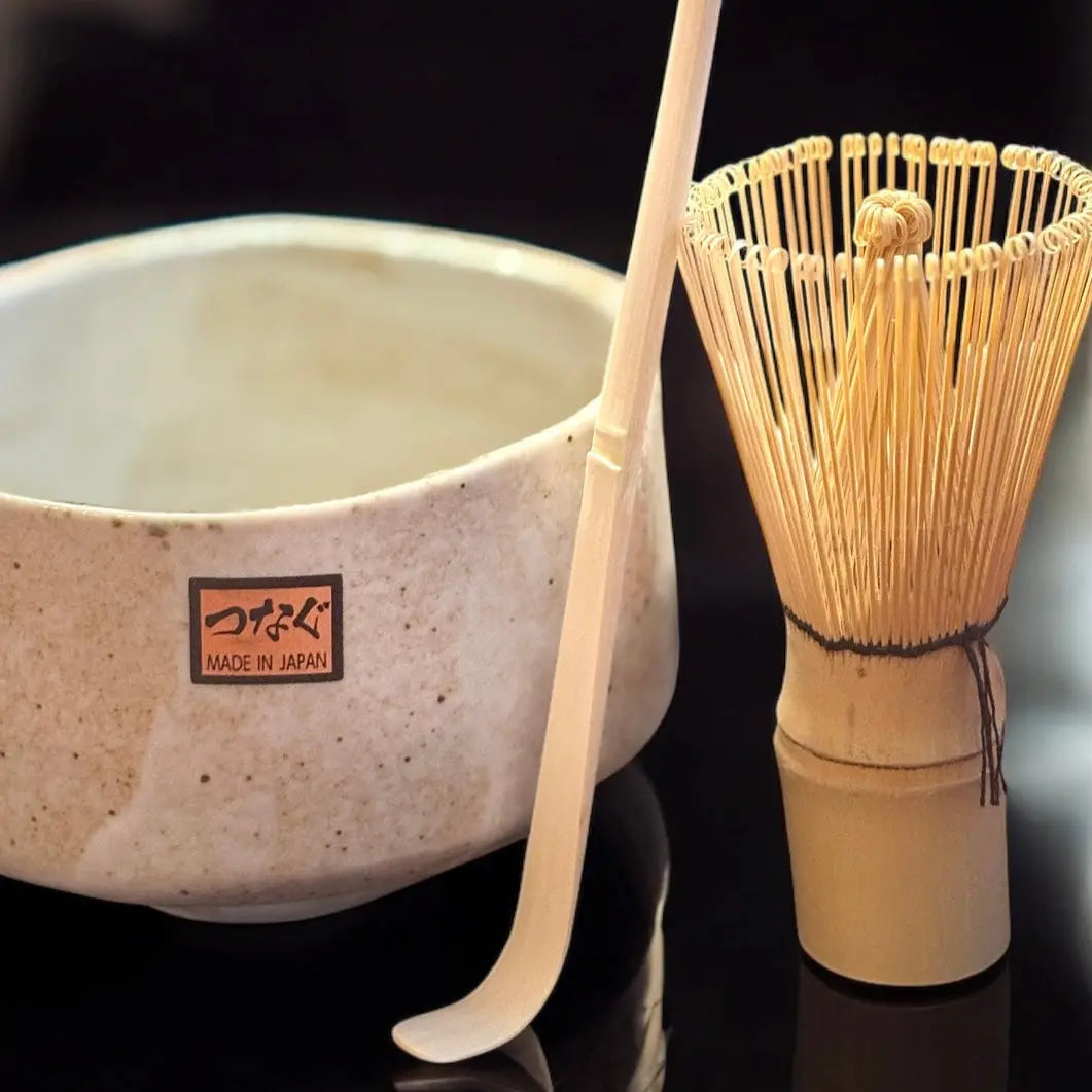 Ein Fitaura Matchabesen Chasen aus weißem Bambus mit 100 Borsten, eine Keramik-Matchaschale und ein Bambuslöffel liegen auf schwarzem Untergrund und erinnern an die japanische Teezeremonie. Die Schale trägt das Label "Made in Japan".