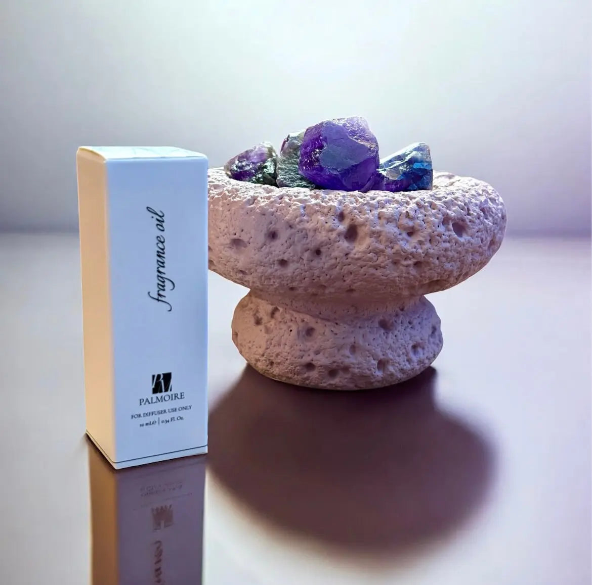 Ein Fitaura Lila Mond Aromatherapie-Diffusor mit Amethyst - Zeder & Adlerholz steht neben einer Box Duftöl; sein strukturierter Steinbehälter mit Amethyststeinen glänzt auf einer reflektierenden Fläche im sanften Licht.
