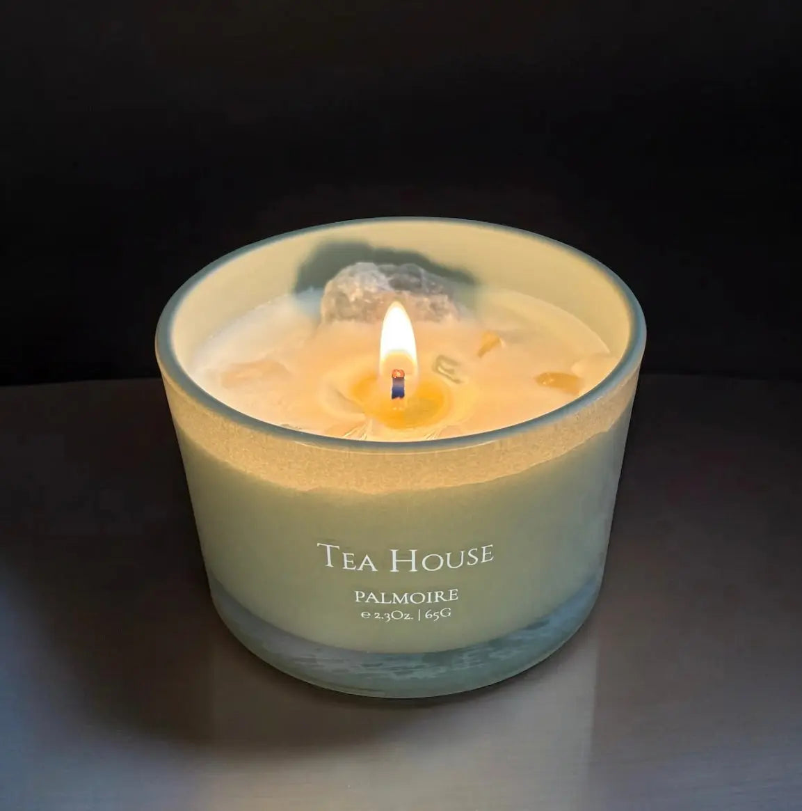 Eine leuchtende Fitaura Tea House Duftkerze mit Bergkristall - Grüner Tee, Jasmin & Zitrone im Milchglas (6.9oz | 195g) sitzt auf einer dunklen Fläche und strahlt Reinheit vor einem schwarzen Hintergrund aus.