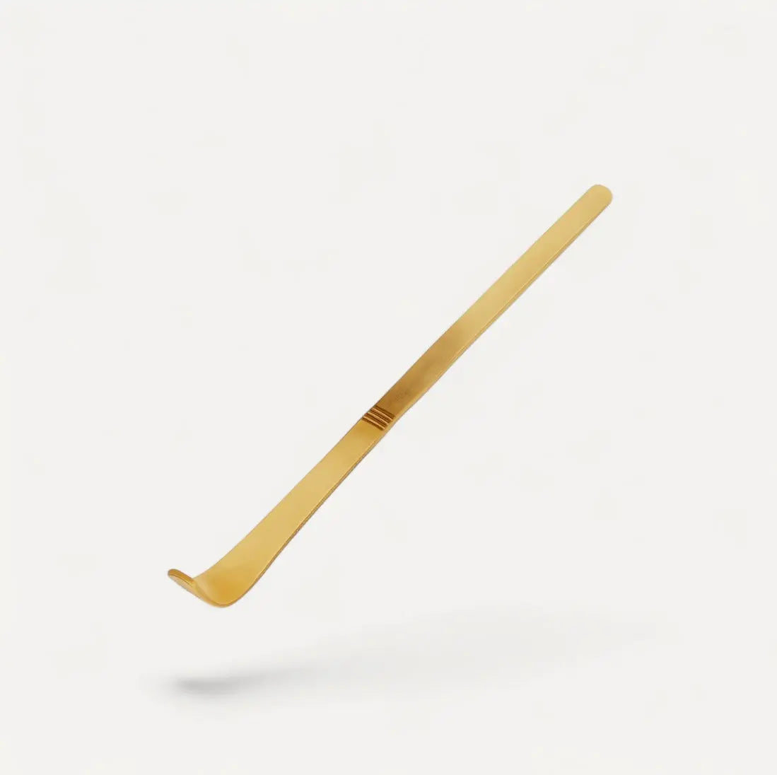 Der Fitaura Matcha Bambuslöffel Chashaku, ein goldfarbener Schuhlöffel aus Metall mit langem Griff und gebogenem Ende, der von traditionellem Design inspiriert ist, schwebt vor einem schlichten weißen Hintergrund.