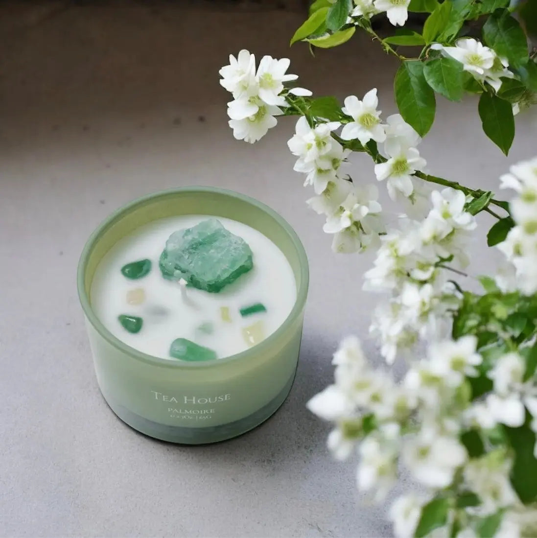 Eine Fitaura Tea House Duftkerze mit Bergkristall - Grüner Tee, Jasmin & Zitrone steht auf einer grauen Fläche neben blühenden weißen Blumen mit grünen Blättern. Die grüne Kerze enthält eingebetteten Bergkristall und weiße Steine.