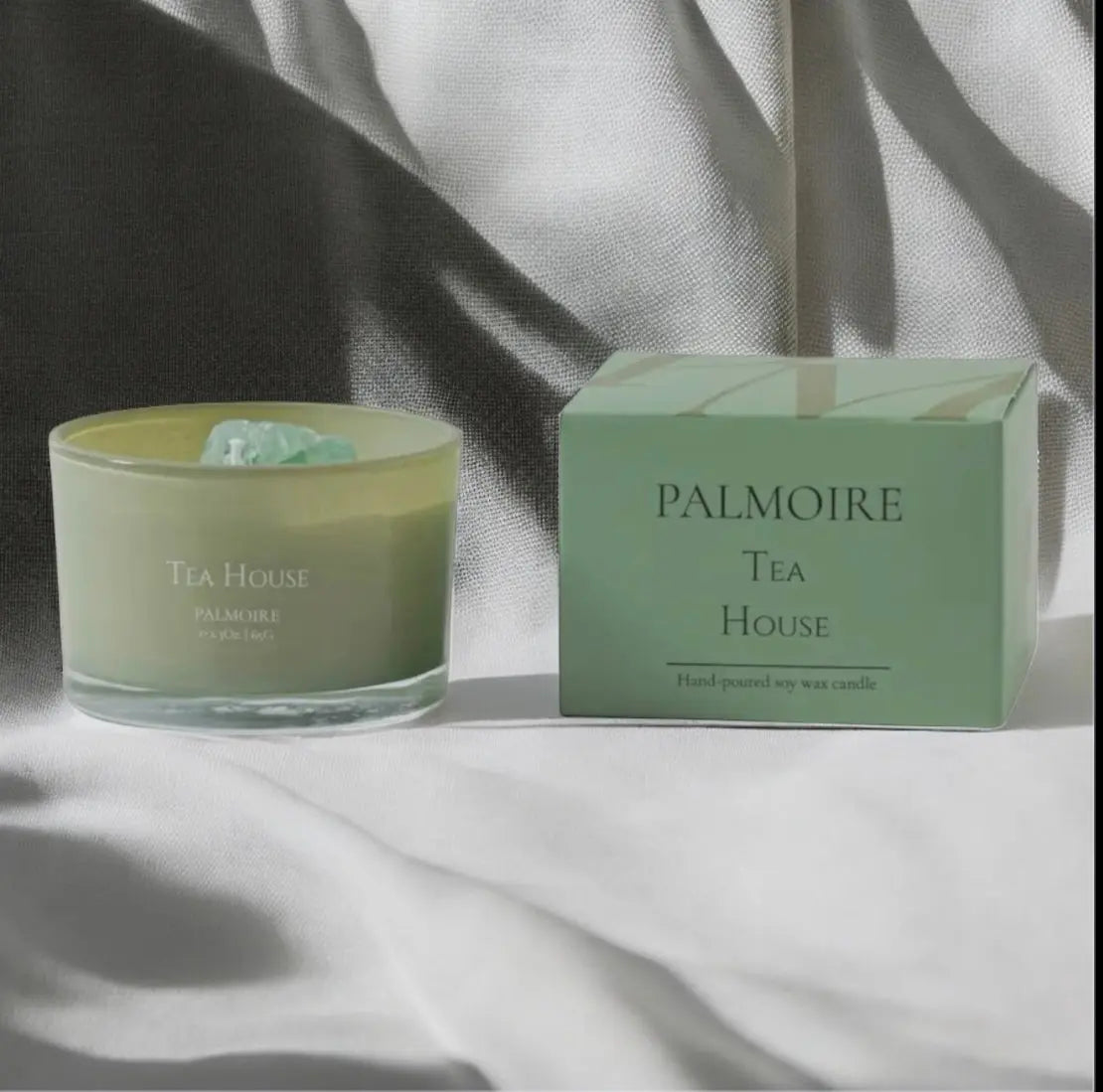 Eine grüne Glas Fitaura Tea House Duftkerze mit Bergkristall - Grüner Tee, Jasmin & Zitrone steht neben der passenden grünen Schachtel auf sonnenbeschienenem Stoff, beide mit minimalistischem Design und Text.