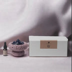 Ein kleiner Fitaura Lila Mond Aromatherapie-Diffusor mit Amethyst - Zeder & Adlerholz und Amethyststeine in einer lavendelfarbenen Schale sitzen neben einer weißen Fitaura-Dose mit beigem Etikett, alles arrangiert auf hellviolettem Stoff.