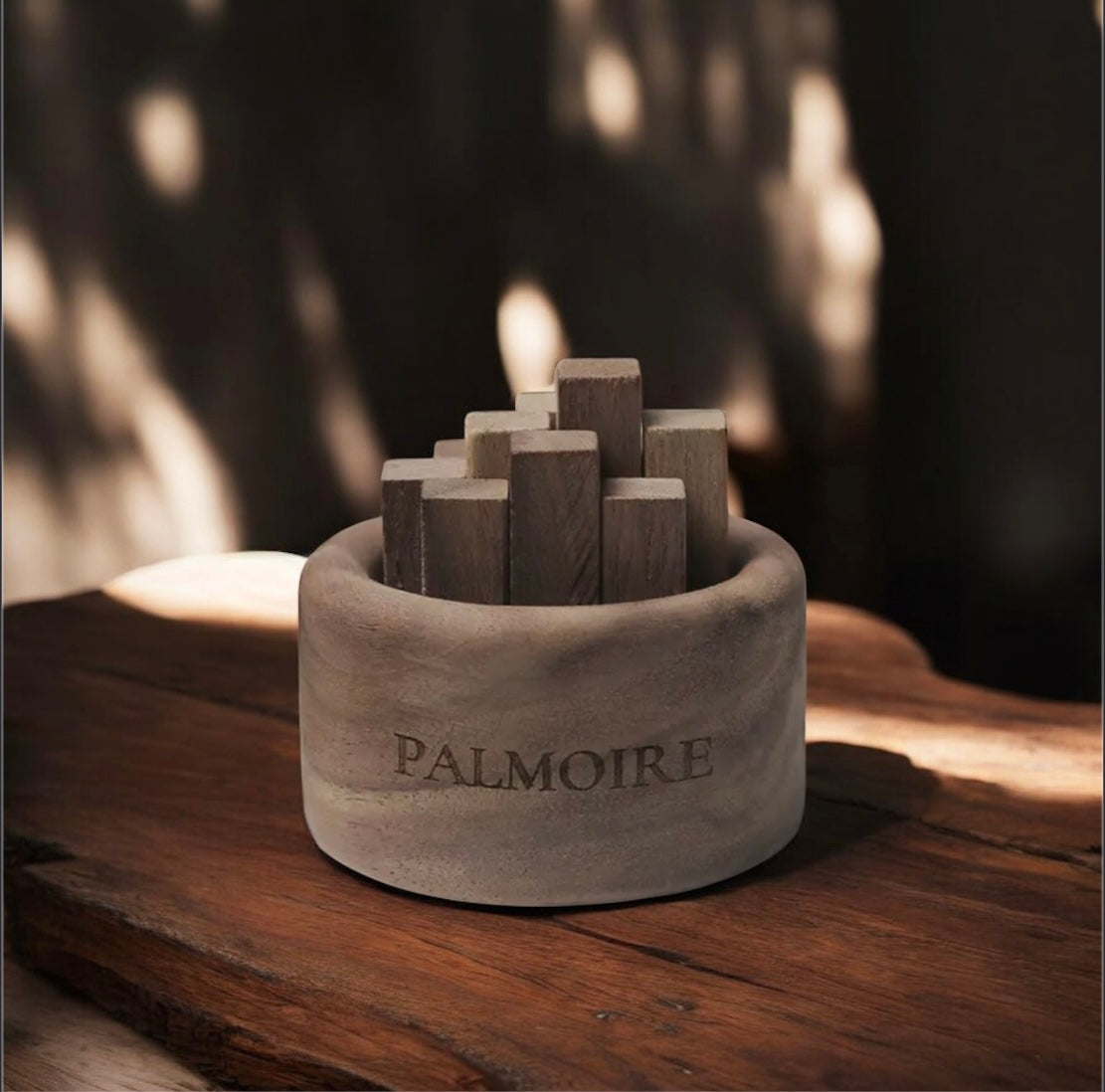 Ein runder Steinbehälter mit der Gravur "PALMOIRE" enthält den Fitaura Mini Holzdiffusor aus Schwarznuss - Jasmin & Rose (To-Go) mit aufrecht stehenden Holzstäbchen, die auf einer rustikalen Holzfläche in schummrigem Sonnenlicht und Schatten stehen.