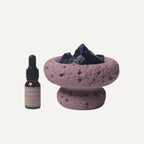 Ein kleines braunes Fitaura-Tropffläschchen steht neben einer mit Amethyst gefüllten lilafarbenen Steinschale, ideal für den Lila Mond Aromatherapie-Diffusor mit Amethyst - Zeder & Adlerholz, alles auf einem schlichten weißen Hintergrund.