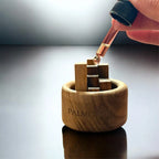 Eine Hand füllt mit einer Pipette ätherisches Öl in den Mini Holzdiffusor aus Schwarznuss - Jasmin & Rose (To-Go) von Fitaura und gibt den Duft auf Holzblöcke im Diffusor für nachhaltige Duftverbreitung auf einer reflektierenden Oberfläche ab.
