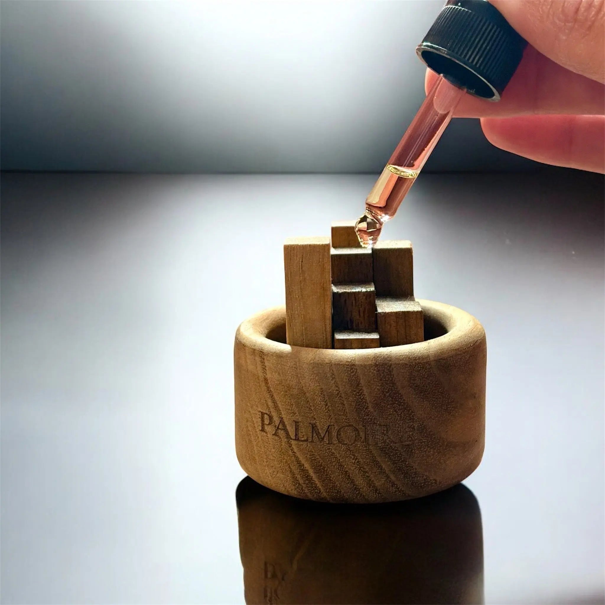 Eine Hand füllt mit einer Pipette ätherisches Öl in den Mini Holzdiffusor aus Schwarznuss - Jasmin & Rose (To-Go) von Fitaura und gibt den Duft auf Holzblöcke im Diffusor für nachhaltige Duftverbreitung auf einer reflektierenden Oberfläche ab.